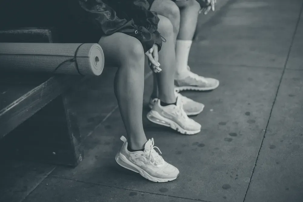 The legs of two people sitting on a wooden bench in running shoes, a rolled-up exercise mat beside them.