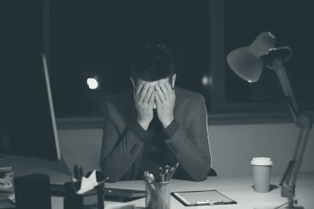 An exhausted man sitting at an office desk at night, his face buried in his hands.