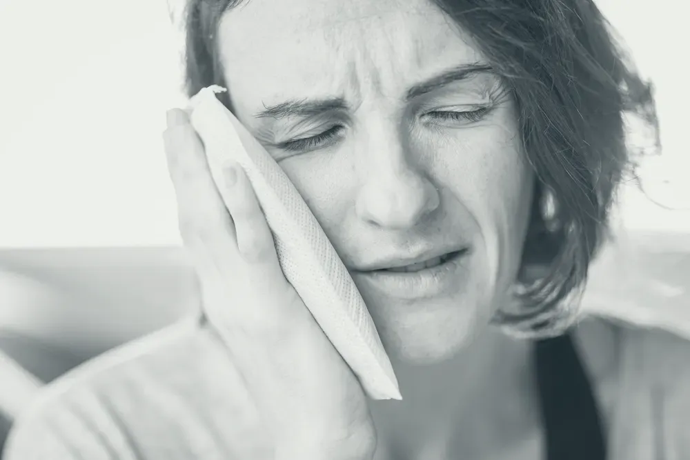 A person holding a cold compress against a swollen cheek indoors, in natural light.