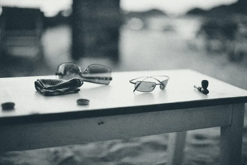 Sunglasses and a pair of prescription glasses resting on a table, in black and white.