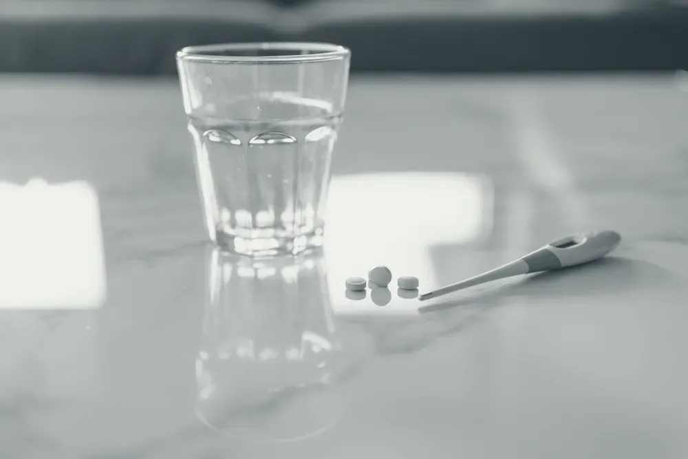 A glass of water, tablets and a thermometer on a light table lit by natural light.