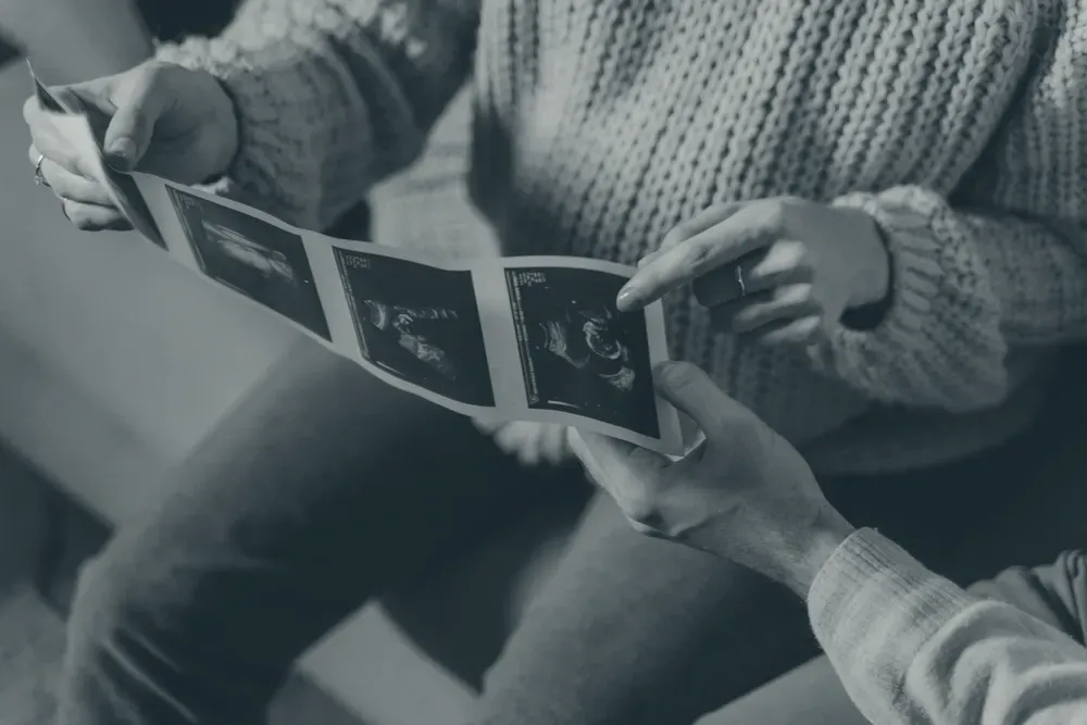 A couple in a cozy room holding and looking at a strip of ultrasound photos, faces out of frame.