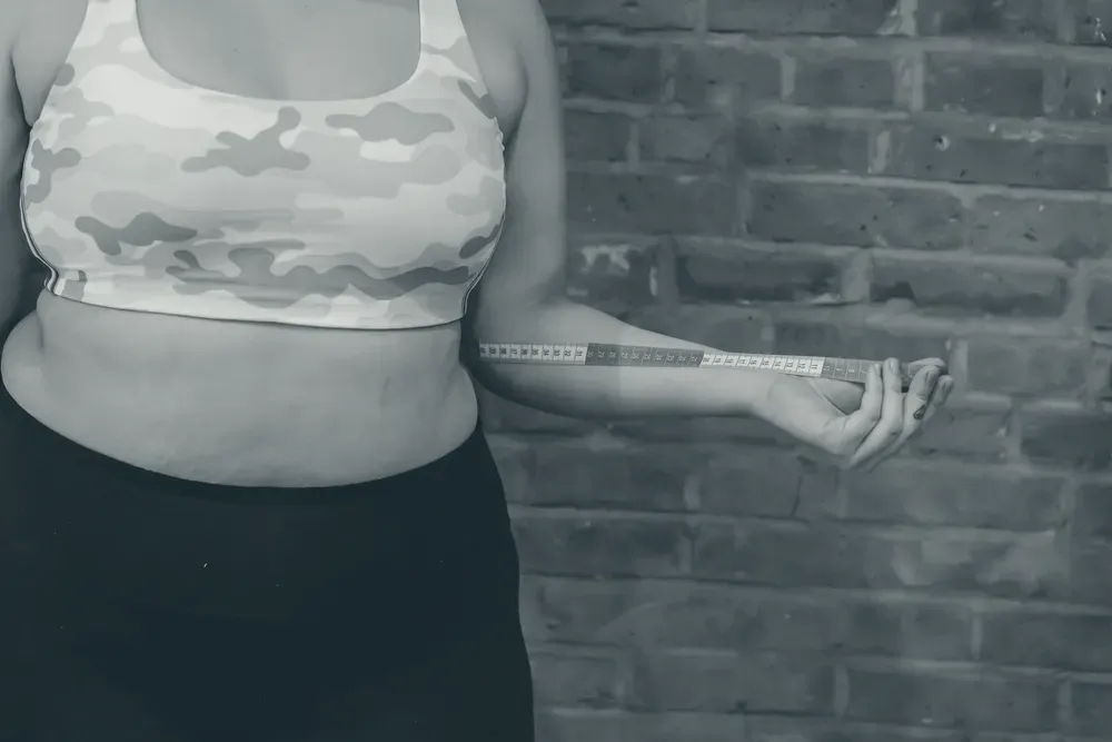 A person measuring their own waist with a red tape measure in front of an exposed brick wall.