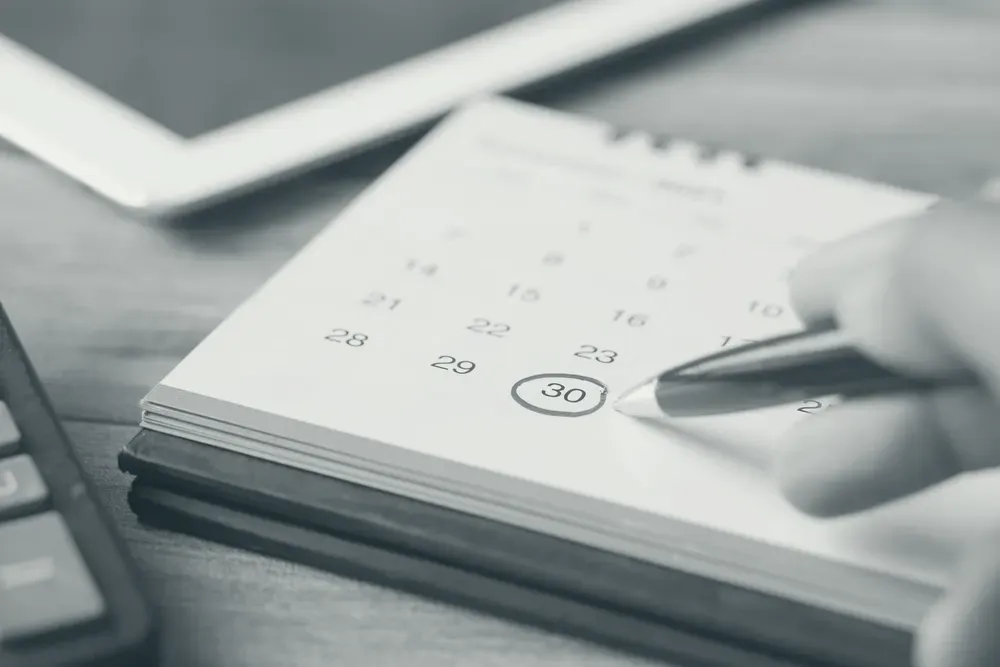 A hand holding a pen over a desk calendar, with one day circled in red.