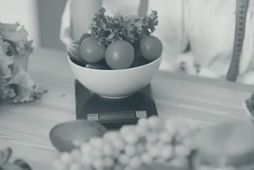 A kitchen scale with a tape measure, a notebook and a bowl of vegetables on a wooden table.