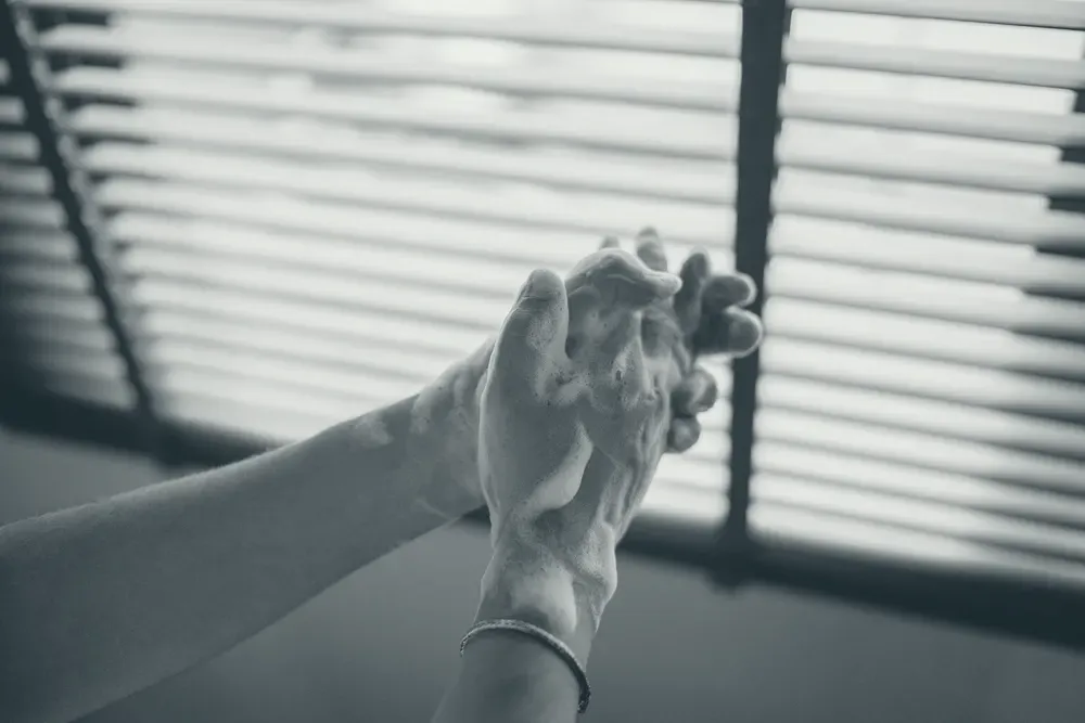 Close-up of soapy hands being washed, with natural light through a blind behind.